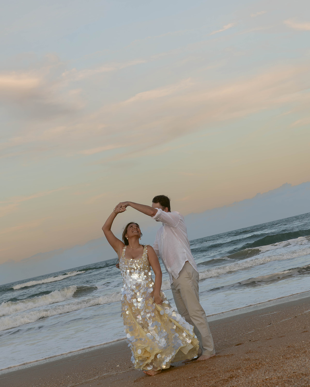 A newlywed couple dances barefoot on the beach at sunset, with the bride wearing a sparkling sequin gown that catches the golden evening light.
