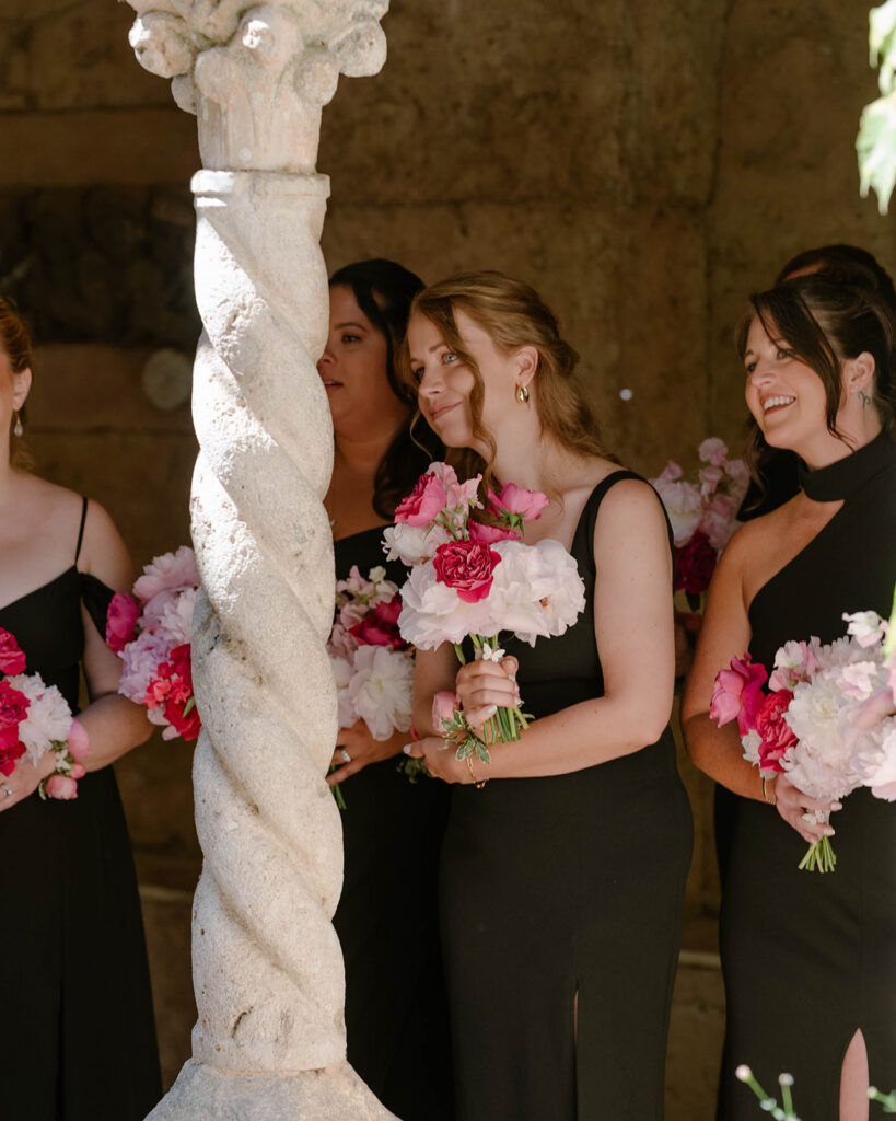 Bridesmaids in black gowns holding pink bouquets beside a marble column