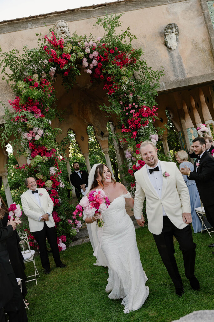 Bride and groom walking arm in arm under a rose-covered arch after their ceremony