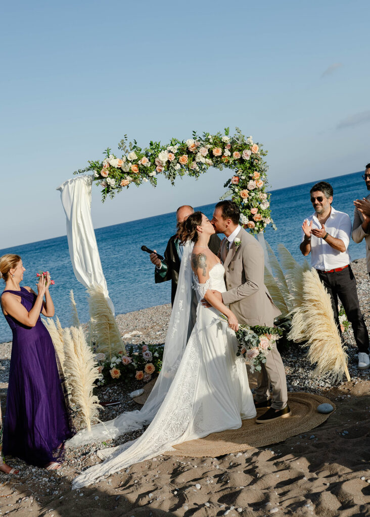 Bride and groom kissing under a floral arch at their Rhodes, Greece beach ceremony