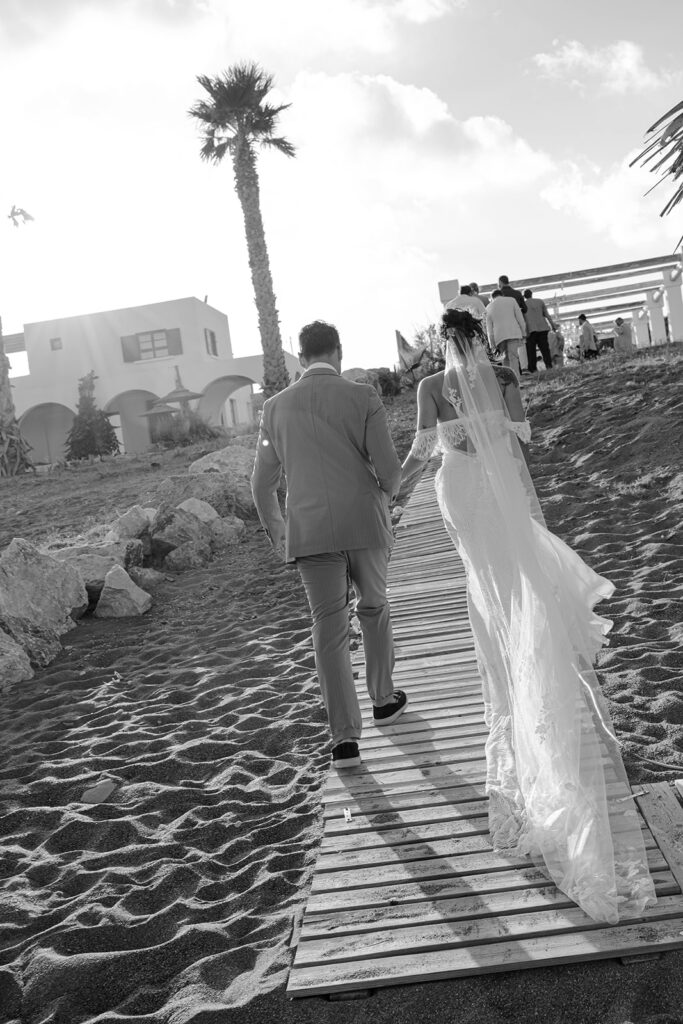 Black and white photo of the groom walking with the bride's veil along a sandy path